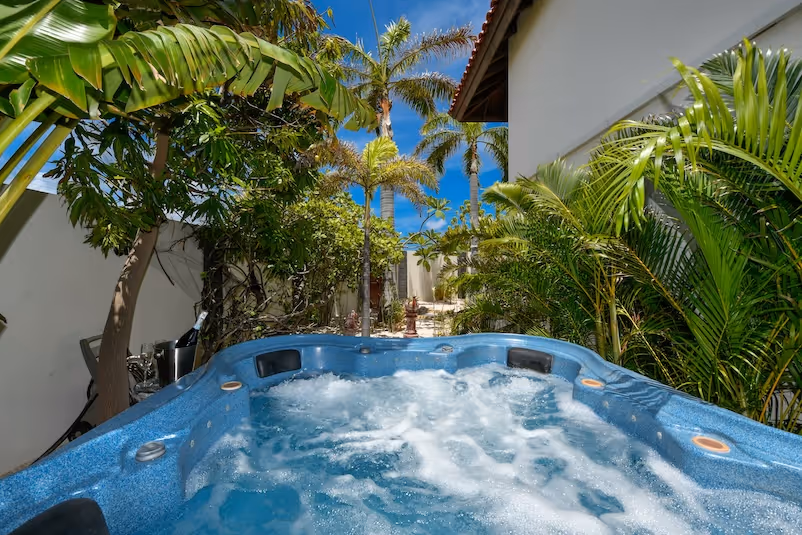 Blue hot tub with bubbling water surrounded by green tropical plants and palm trees under a clear blue sky.