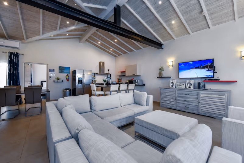Modern open-plan living room with a large gray sectional sofa, ottoman, wall-mounted TV, and dining area with six chairs near the kitchen.