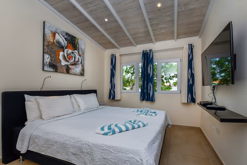 Minimalist bedroom with a large bed covered in white bedding, two folded striped towels, blue and white curtains, a wall-mounted TV, and a floral painting above the headboard.
