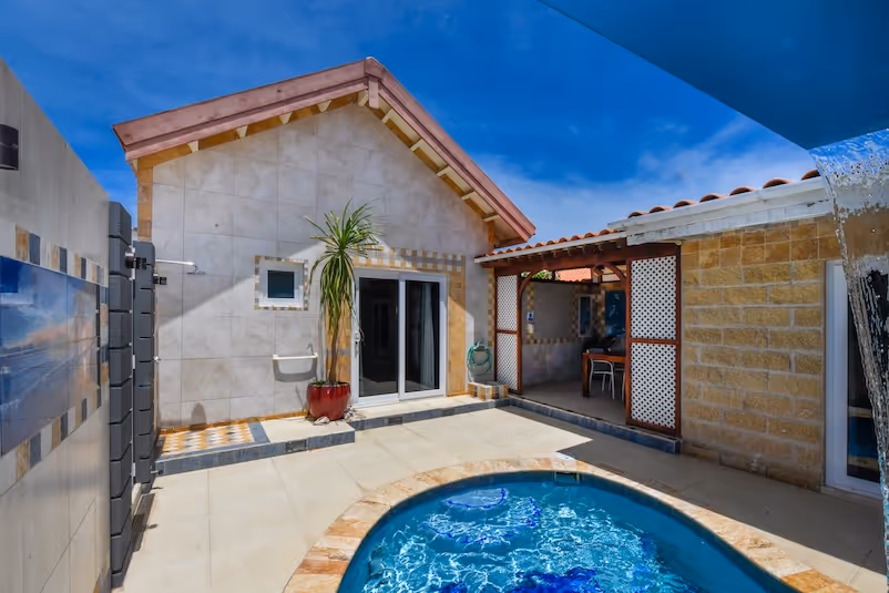 Small outdoor pool with water cascading from a spout next to a tiled villa featuring sliding glass doors and a potted plant under a clear blue sky.