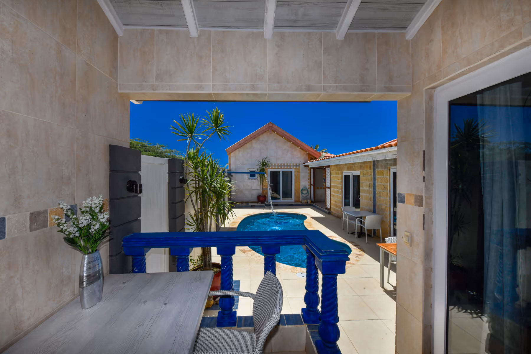 View from a covered patio with a table and chair overlooking a small outdoor pool surrounded by beige tiled buildings under a clear blue sky.