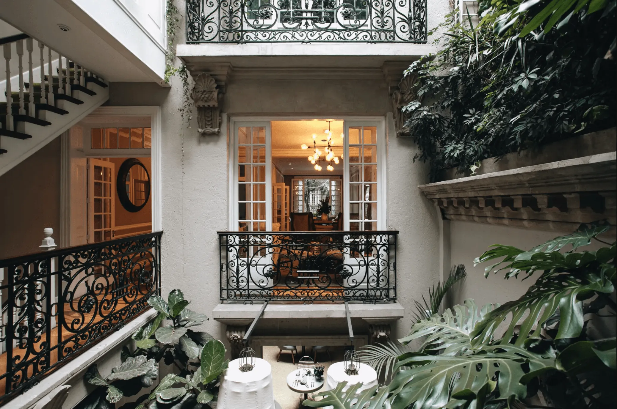 Indoor courtyard with lush green plants, ornate black wrought iron railings, a small table with two white stools, and large windows showing a warmly lit interior.