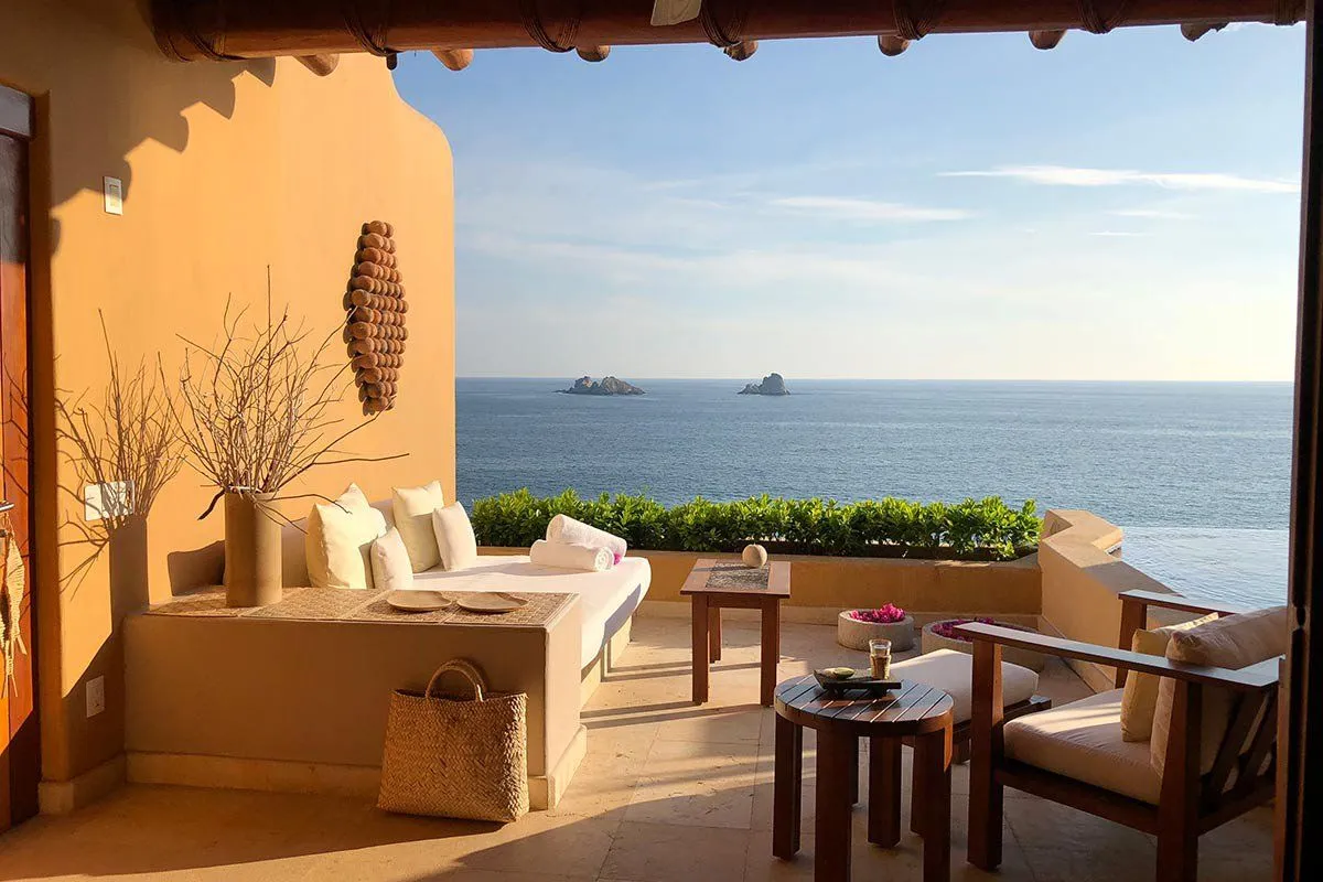 Sunny balcony with cushioned seating, wooden tables, and a sea view with two small rocky islands.