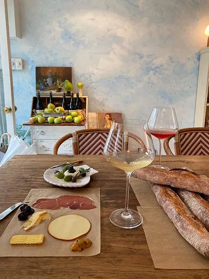 Table set with sliced cheese, cured meats, baguettes, olives, and two glasses of wine in a cozy dining space with a shelf of bottles and green apples in the background.