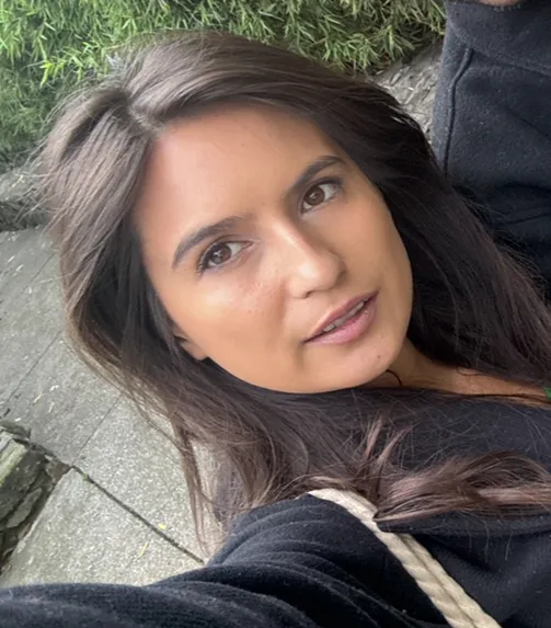 Young woman with long dark hair wearing a black top, taking a selfie outdoors near green plants and a stone path.