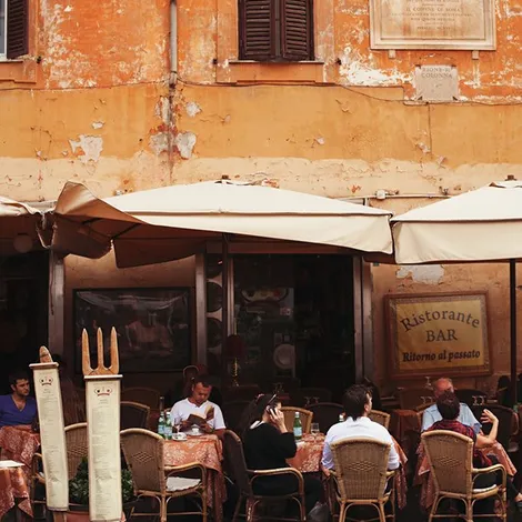 Outdoor seating area of a rustic Italian restaurant with patrons dining under beige umbrellas in front of a weathered orange building.