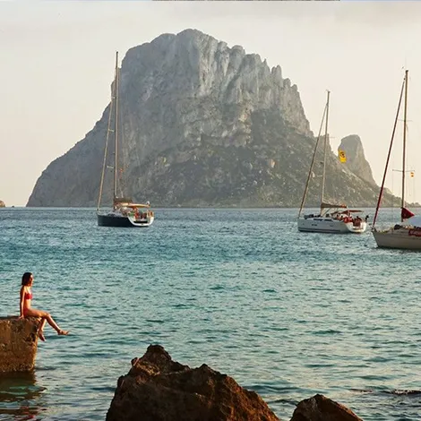 Woman sitting on a rock by the sea with sailboats on calm water and a large rocky island in the background during daylight.