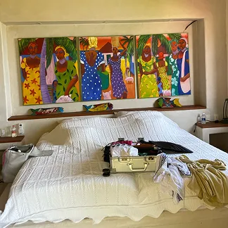 Bedroom with a large white bed covered in a textured blanket, an open suitcase, and clothes on it, and a colorful painting of women on the wall above the bed.