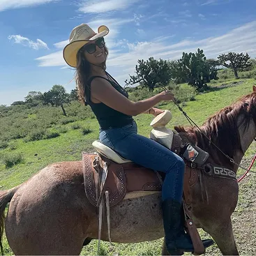 Woman wearing a cowboy hat, sunglasses, black tank top, and jeans sitting on a horse in a grassy field with trees under a partly cloudy sky.
