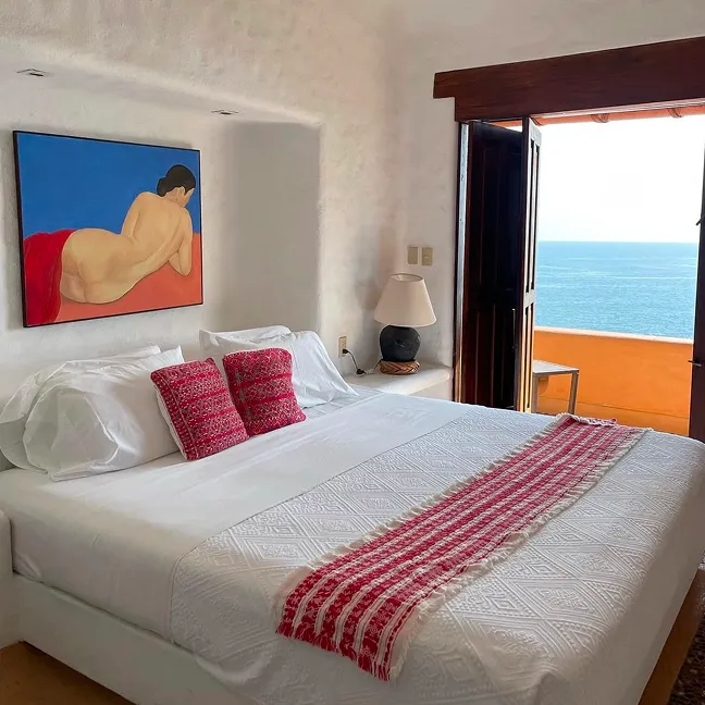 Bedroom with a white bedspread, two red patterned pillows, a red runner, a painting of a woman on the wall, and an open door revealing an ocean view.