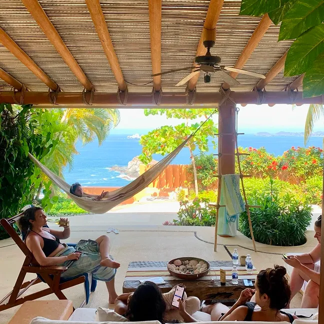 Group of people relaxing on chairs and a hammock under a wooden pergola overlooking a tropical ocean view with lush greenery.
