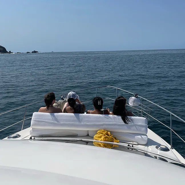 Four people sitting on a white cushioned bench at the front of a boat, facing calm ocean waters under a clear blue sky.