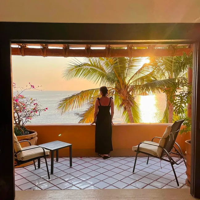 Woman in a black dress standing on a balcony overlooking palm trees and the ocean at sunset.