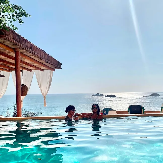 Two women relaxing and smiling in an infinity pool overlooking a sunny ocean with small rocky islands in the distance.
