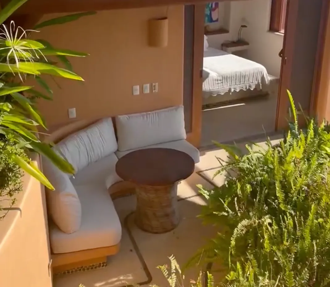 Outdoor patio area with a curved cushioned sofa, round wicker table, green plants, and a view into a bedroom with a white bedspread.