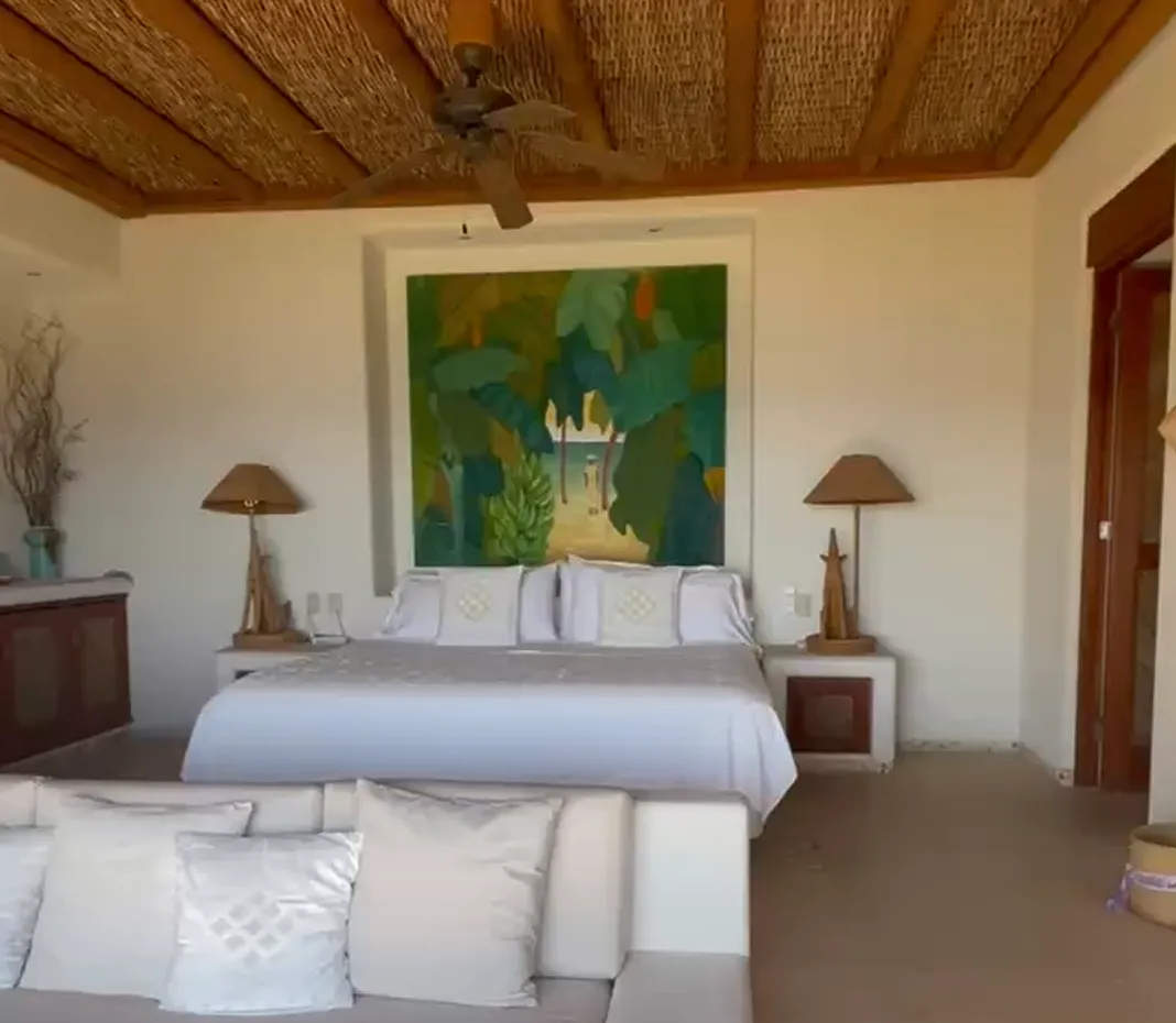Cozy bedroom with a large bed, white linens, two wooden bedside lamps, a tropical-themed painting above the bed, and a beige couch with cushions in the foreground.