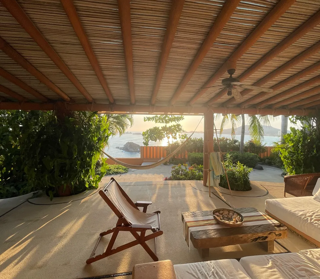 Covered patio with wooden ceiling, hammock, outdoor seating, and a view of the ocean and tropical plants at sunset.