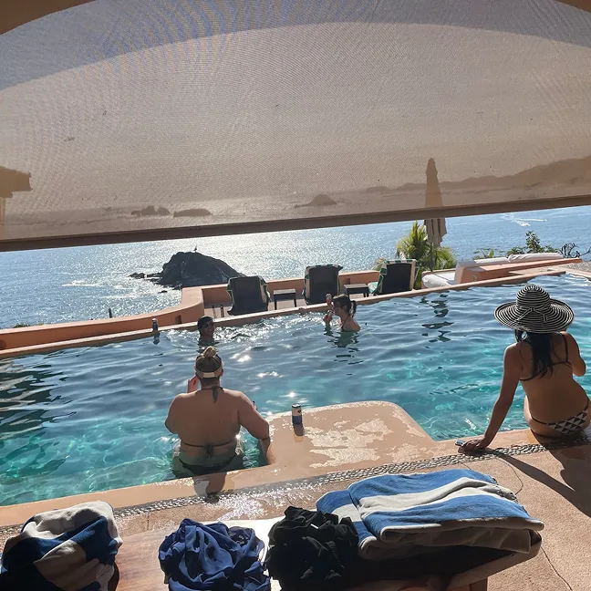 Four people relaxing in and around an infinity pool overlooking a sunlit ocean and rocky coastline under a large shade canopy.