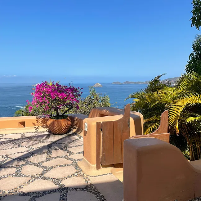 Terrace with stone and pebble flooring, a potted pink bougainvillea plant, tropical palm plants, and a sea view with islands under a clear blue sky.