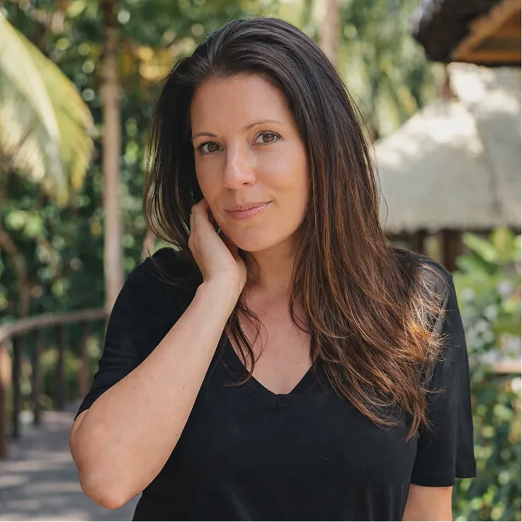 Woman with long brown hair wearing a black V-neck shirt, standing outdoors with greenery and a thatched roof structure in the background.