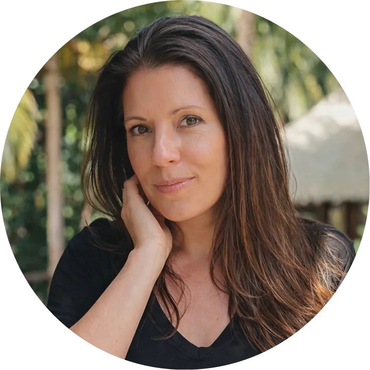Woman with long brown hair wearing a black shirt, touching her neck, with blurred greenery and a hut in the background.