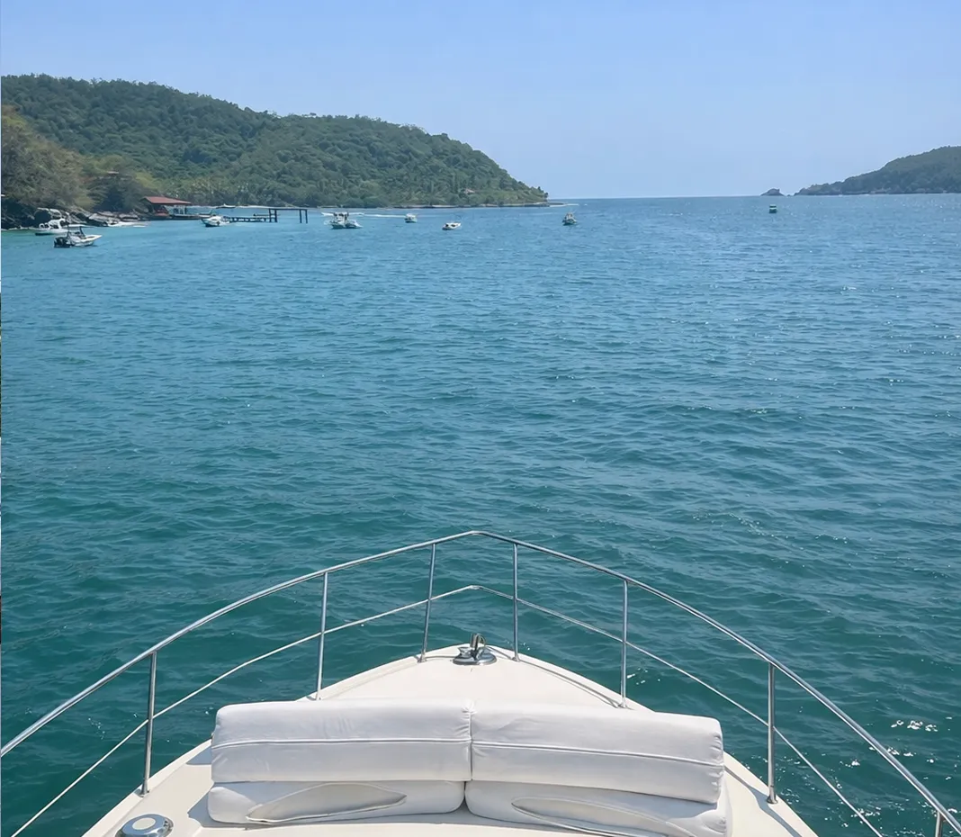 View from the bow of a boat looking out over calm blue sea with small boats near green, forested hills under a clear sky.