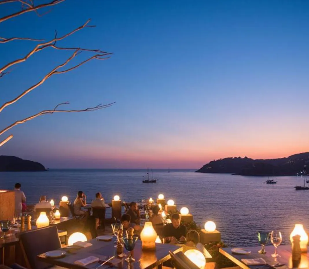 Outdoor restaurant terrace at sunset overlooking calm sea with sailboats and distant hills.