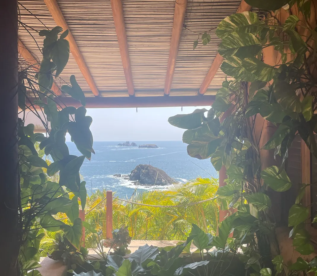 View of ocean with small rocky islands framed by lush green plants and a bamboo roof overhead.