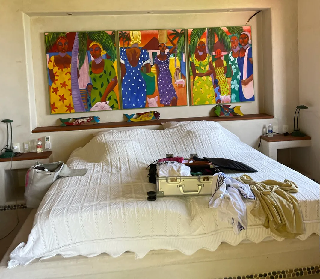 Bedroom with a large white bedspread-covered bed, colorful tropical paintings of women above headboard, and personal items including a silver suitcase and clothes on the bed.
