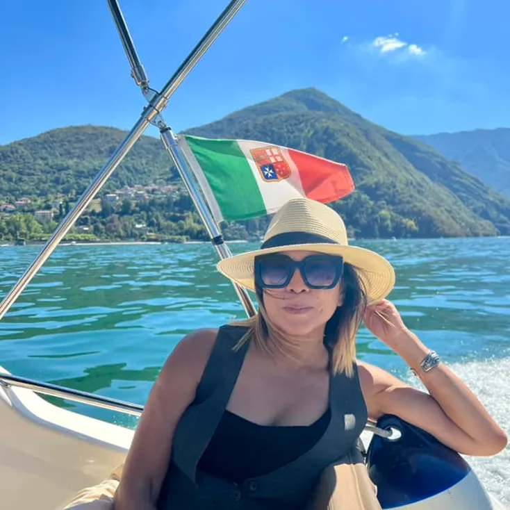 Woman wearing a hat and sunglasses relaxing on a boat with an Italian flag and green mountains in the background.