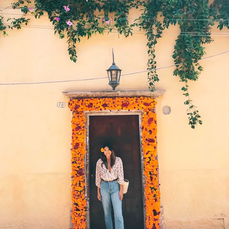 Woman with a flower in her hair standing in a doorway framed by vibrant orange marigold decorations, with green vines hanging above.