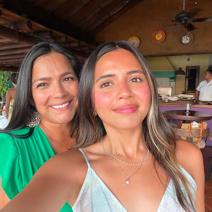 Two smiling women with long dark hair taking a selfie indoors at a restaurant with wooden ceiling and round tables.