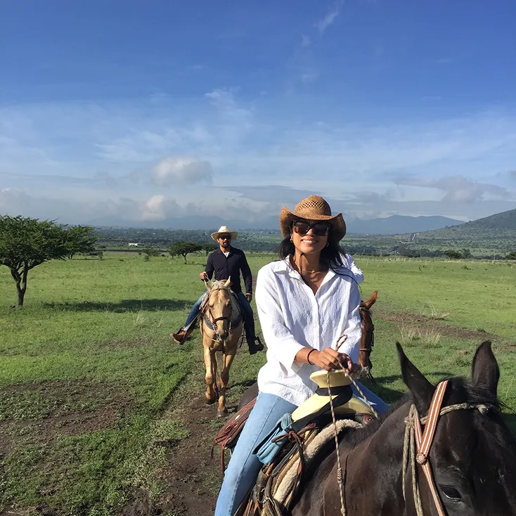 A smiling woman wearing a cowboy hat and sunglasses rides a dark horse on a grassy trail with a man on a light brown horse behind her under a blue sky.