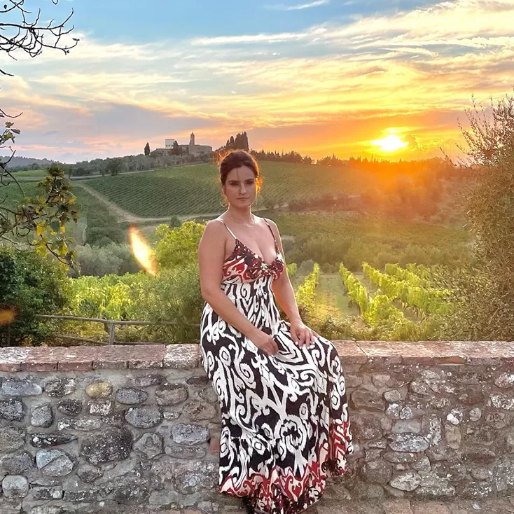 Woman in a patterned dress sitting on a stone wall with a vineyard and sunset in the background.