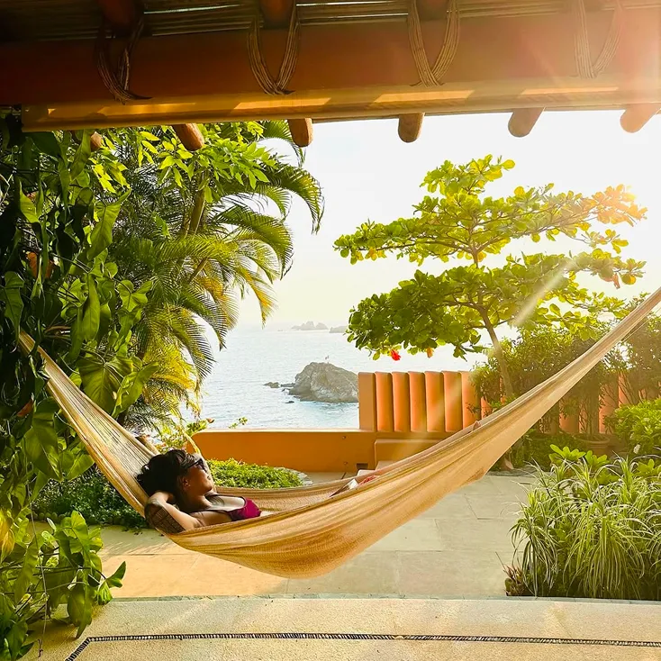Person relaxing in a hammock on a porch surrounded by tropical plants with a view of the ocean and rocks in the distance at sunset.