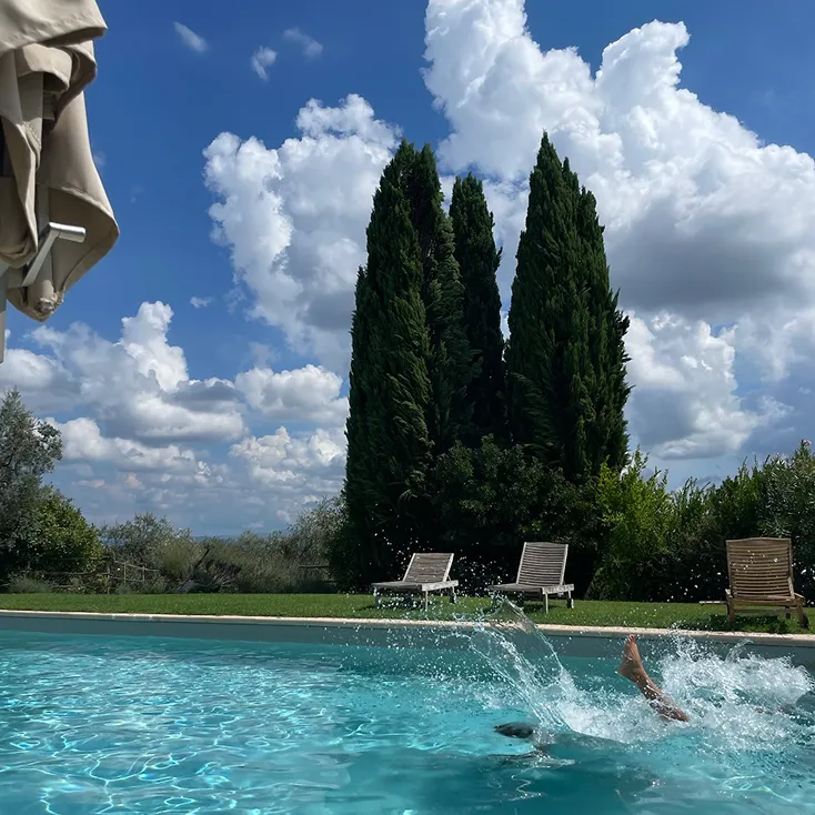 Person splashing into a clear blue swimming pool with lounge chairs and tall cypress trees under a partly cloudy sky.