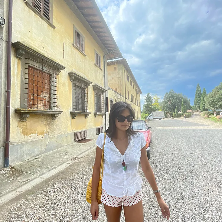 Woman wearing sunglasses, white top, and polka dot shorts walking on a gravel pathway next to an old yellow building under a cloudy sky.