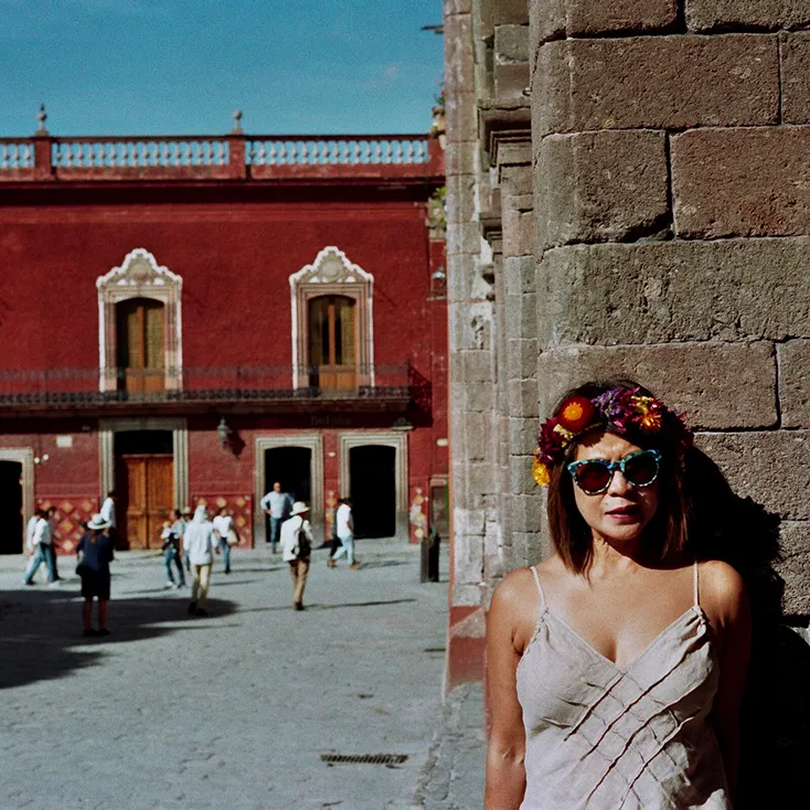Woman wearing sunglasses and a colorful flower crown standing against a stone wall with a red historic building and people walking in the background.
