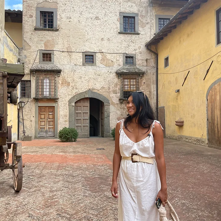 Woman in a white dress smiling and standing in a rustic courtyard with old stone and yellow plaster buildings.