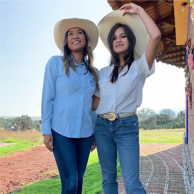 Two women wearing white cowboy hats, one in a light blue shirt and the other in a white shirt, standing outside near a building with a tiled roof and green grass in the background.