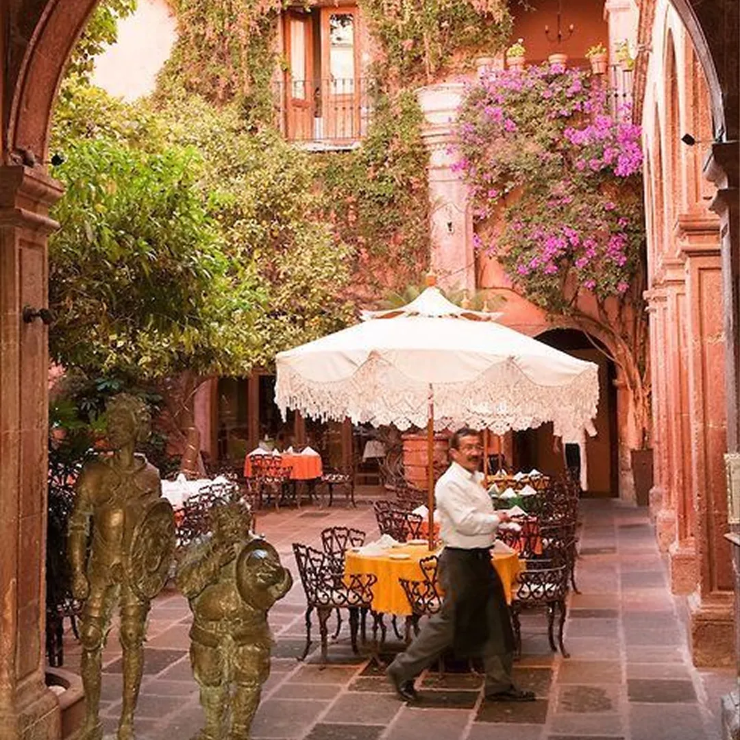 Outdoor courtyard restaurant with wrought iron tables covered by umbrellas and a waiter walking by, surrounded by plants and historic architecture.