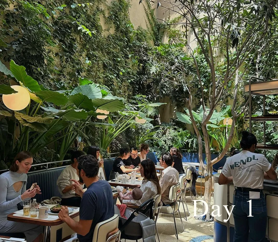 Indoor café with lush green plants and people sitting at tables enjoying food and drinks, with a server wearing a MENDL Delicatessen shirt.