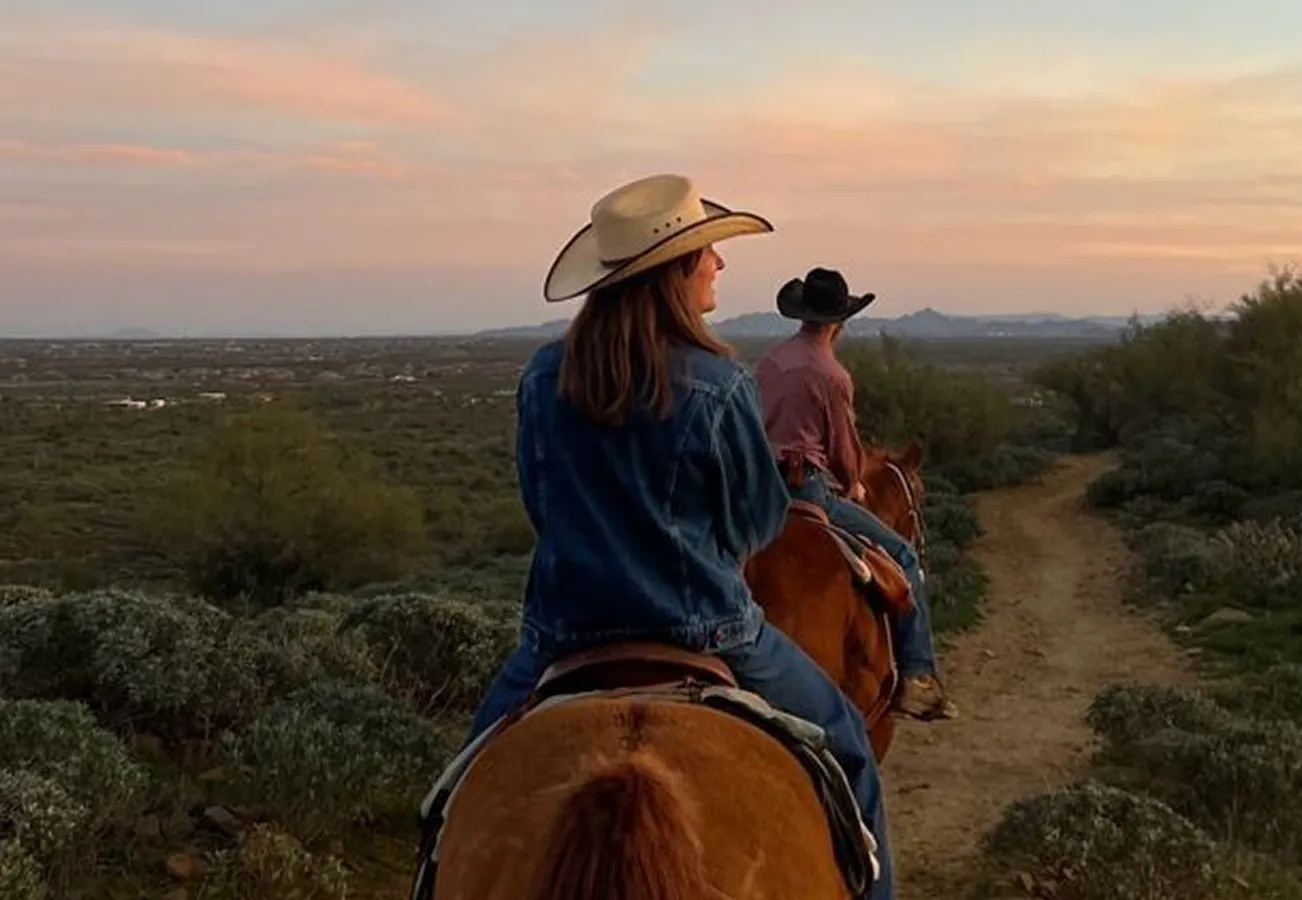 Two people wearing cowboy hats riding horses on a dirt trail through a desert landscape at sunset.