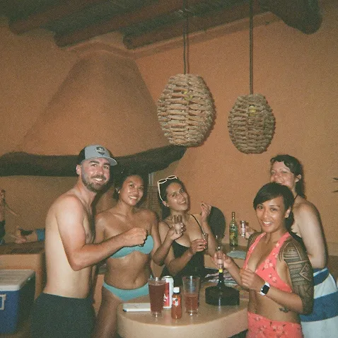 Group of five people in swimwear smiling and holding drinks around a small table indoors with hanging wicker lights above.