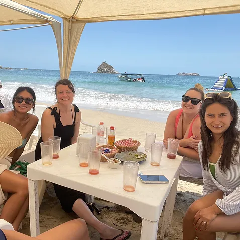 Group of five women sitting around a white table on a beach under a canopy with drink cups and snacks, ocean and small islands visible in the background.