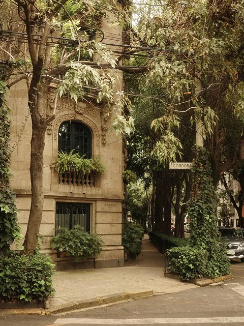 Corner of a stone building with arched windows and abundant green foliage on trees and plants along a sidewalk beneath.