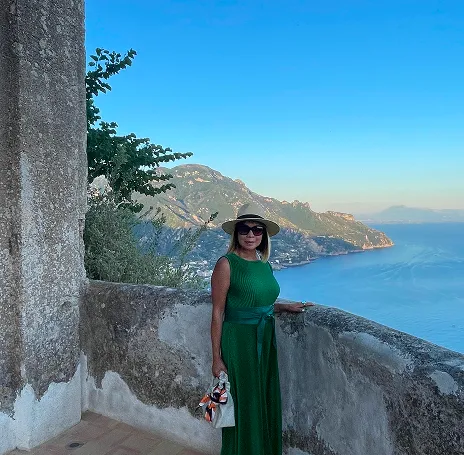 Woman in a green sleeveless dress and hat standing by a stone railing overlooking a mountainous coastline and sea under a clear blue sky.