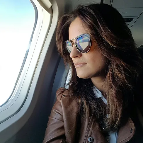Woman wearing sunglasses and a brown leather jacket looking out of an airplane window.