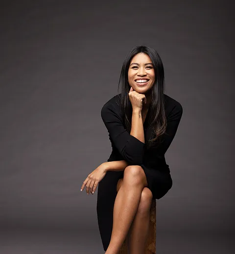 Smiling woman with long dark hair sitting cross-legged on a stool against a dark background.