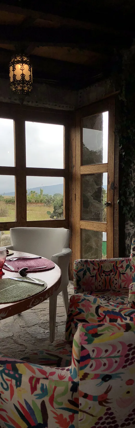 Cozy indoor dining area with colorful floral-patterned chairs, a round wooden table set with placemats, and large windows overlooking a green landscape.